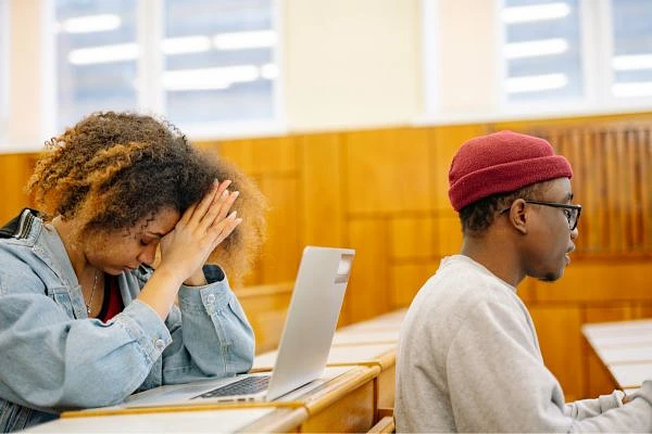 Two people are sitting at desks in a classroom, with one looking stressed and the other focused on their computer.
