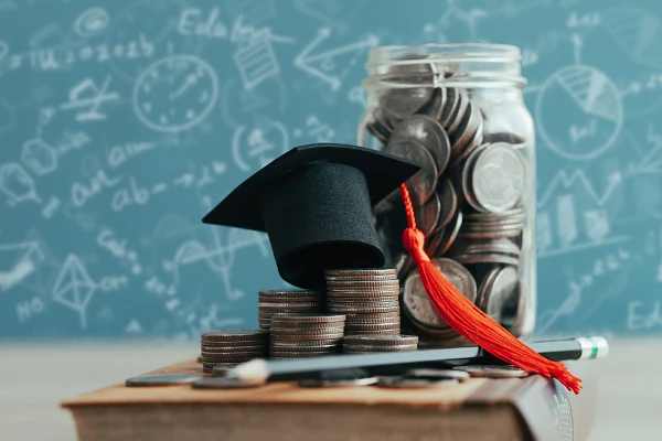 A graduation cap sits atop stacks of coins on a book, with a jar of coins and a chalkboard with mathematical drawings in the background.